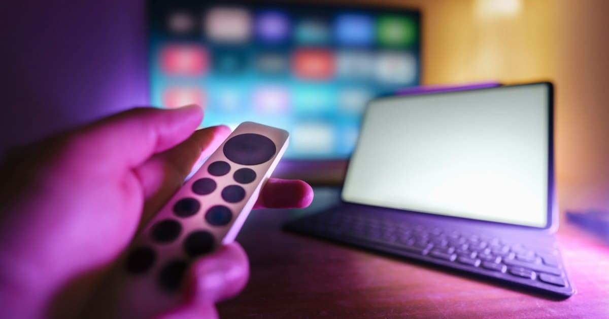 Hand holding a streaming remote in front of a TV with a blurred on-screen menu grid, next to a tablet with a keyboard
